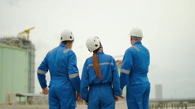 Three professional industrial engineers in blue coveralls and white hard hats discuss and point at a large storage tank facility. Factory workers inspecting plant operations and site progress.