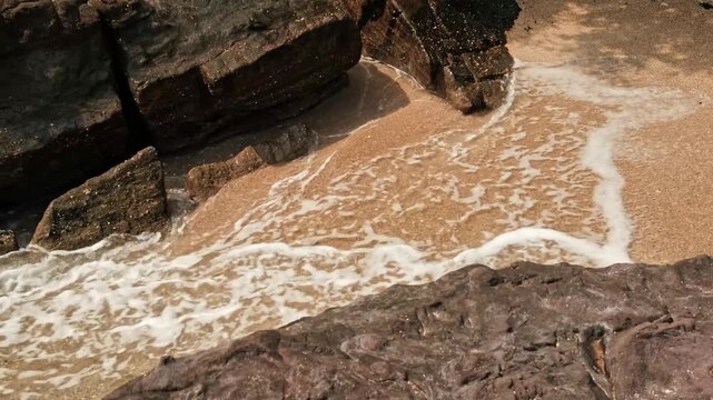 From an elevated perspective, ocean water rushes onto a small sandy cove, breaking into white foam against dark stone geological formations.