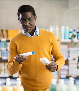 Focused African American in casual ochre jumper and glasses examining healing ointment box in pharmacy, checking instructions before using for wounds or burns