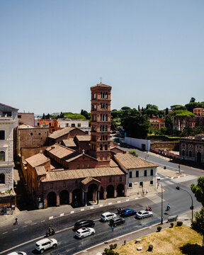 Aerial view of the Basilica of Santi Giovanni e Paolo with its distinctive brick bell tower, surrounded by historic buildings and a busy street in Rome, Lazio, Italy.
