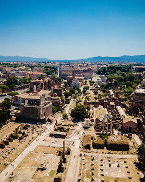Aerial view of the Roman Forum and the Colosseum surrounded by ancient ruins and lush trees under a clear blue sky in Rome, Lazio, Italy.