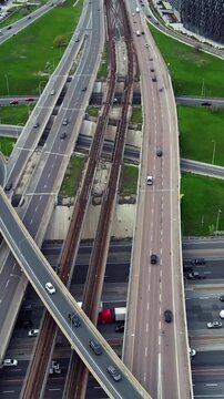 Aerial view of highway interchange traffic with curving elevated ramps, rail lines, and moving cars through urban infrastructure, surrounded by green medians and adjacent roads in city network