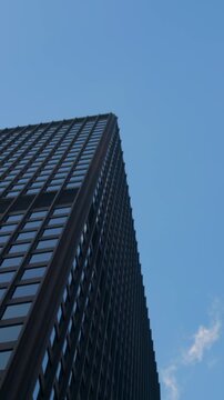 Modern skyscraper rises against blue sky, seen from low angle with dark glass facade, repeating window grid, and sharp geometric lines, creating urban architecture composition with vertical symmetry.