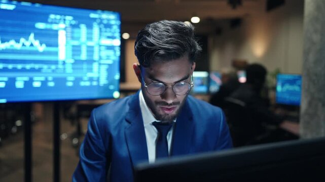 Front view male broker wearing glasses and blue suit while looking at computer screen. Trader analyzing financial charts in dark office. Businessman focusing on stock market data late at night.