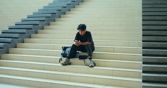 Young man waiting on wide staircase while checking smartphone, seated beside backpack on empty steps in minimal interior with repeating stair pattern and clean architectural lines.