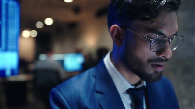 Close up serious male broker wearing glasses and blue suit while looking at computer screen. Trader analyzing financial charts in dark office. Businessman focusing on stock market data at night.