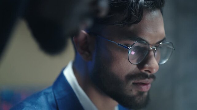 Close up of handsome businessman in glasses and looking at computer screen. Male trader analyzing financial data in dark office. Focused broker working with stock market and listening to colleague.