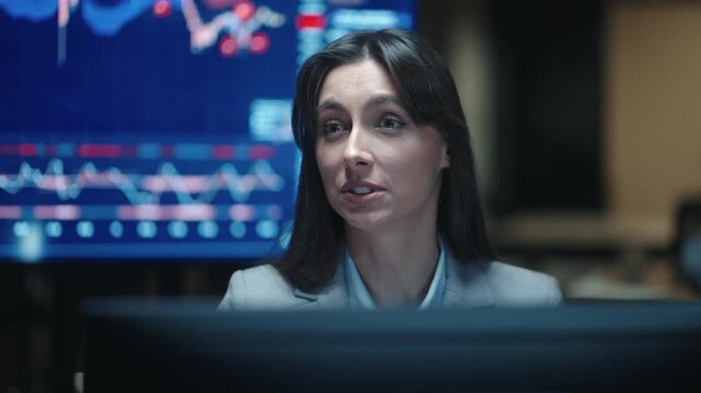 Frustrated Caucasian female broker laughing while talking at computer near financial charts. Woman trader coping with stock market stress in dark office. Taking pause during night work.