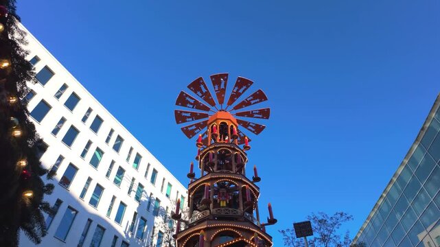 Traditional wooden german christmas pyramid with nativity scenes spinning slowly in a modern city square during the festive holiday season against a clear blue sky and contemporary buildings