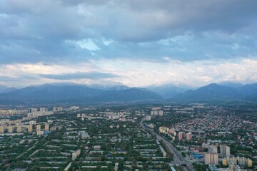 Fototapeta premium Aerial view of sprawling urban cityscape nestled against snow-capped mountain ranges under dramatic cloudy skies, Almaty, Kazakhstan