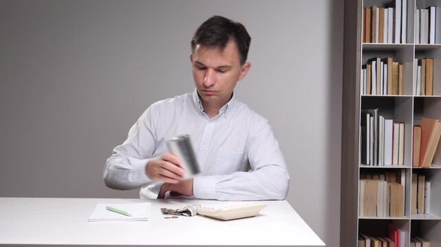 Man is pouring a few coins from an empty metal container onto a white table, showing financial difficulties, poverty, and saving money during a crisis