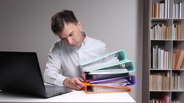 Man concentrating at his desk, working with a laptop while compiling a large stack of colorful binders filled with important files, documents, and paperwork in an organized office setting