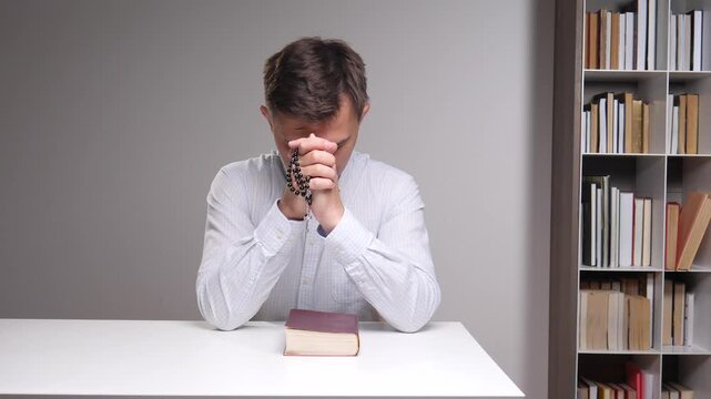 Person is holding a rosary and bowing head in deep prayer, hands clasped over a bible on a white table with a bookshelf visible in the background, expressing faith and spiritual devotion