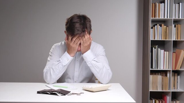 Man in a light shirt holding his head in his hands, feeling immense stress and frustration over financial problems, with an empty wallet and coins visible on the table