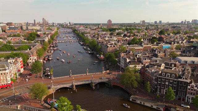 Drone orbits above Amsterdam Amstel river with boats and a historic bridge flanked by classic canal house facades