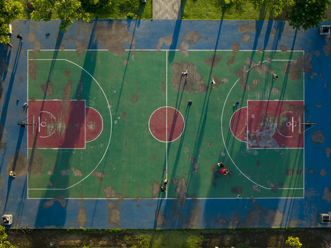 Aerial view of Alun-Alun Bangli basketball court with worn green and red surface, long shadows, and people playing in Kecamatan Bangli, Bali, Indonesia.