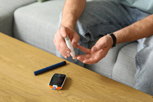 Diabetic young man using lancet pen on sofa at home, closeup