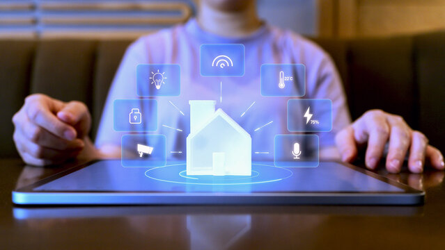 A person using a tablet to control a smart home system with digital hologram icons for lighting, security, energy, and IoT connectivity, representing modern home automation technology.