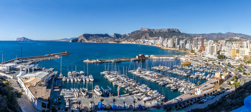 Panoramic view of Calpe, Alicante, from the ascent to Pe&ntilde;&oacute;n de Ifach, overlooking the Mediterranean coastline, marina, sandy beaches, and the surrounding mountains under a clear blue sky.