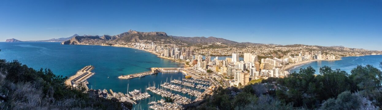 Panoramic view of Calpe, Alicante, from the ascent to Pe&ntilde;&oacute;n de Ifach, overlooking the Mediterranean coastline, marina, sandy beaches, and the surrounding mountains under a clear blue sky.