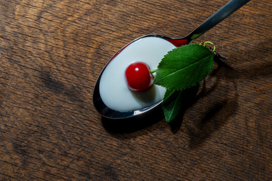 Fresh red cherry on white yogurt in metal spoon. Top view single ripe cherry with green leaves on creamy white yogurt in metal spoon over dark rustic wooden background representing healthy snack