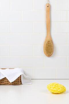 Minimalist bathroom mockup with a long handle wooden body brush hanging on a white subway tile wall. Yellow sea sponge and a white cotton towel in a wicker basket on the counter.