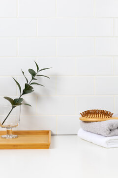 Minimalist bathroom product mockup with white subway tile background. Wooden tray with green leaf in glass and stacked towels with hairbrush for cosmetic display.