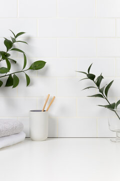 Eco-friendly dental care mockup background with white subway tile wall. Bamboo toothbrushes in a ceramic cup surrounded by fresh green leaves and soft folded towels.