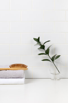 Minimalist bathroom product mockup with white subway tile background. Wooden tray with green leaf in glass and stacked towels with hairbrush for cosmetic display.