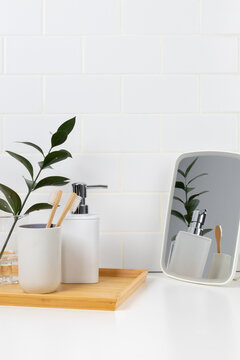 Eco-friendly oral care and bathroom amenities mockup. Bamboo toothbrushes, white soap dispenser on a wooden tray, and tabletop mirror with reflection against white subway tile wall.