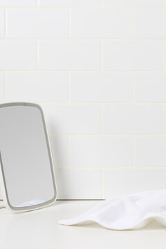 Minimalist beauty and skincare routine mockup background. White tabletop mirror and soft cotton towel on a counter against white subway tile wall with copy space.