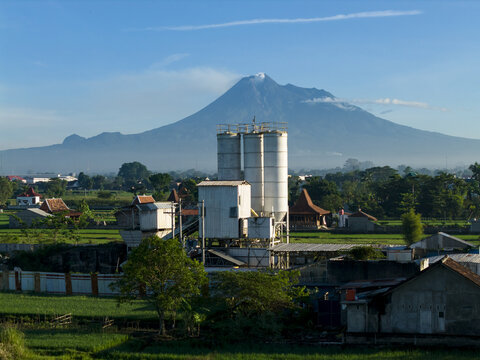Aerial view of concrete production site with tall silos and industrial equipment, surrounded by green rice fields, with a volcano in the background in Kecamatan Prambanan, Jawa Tengah, Indonesia.