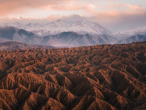 Aerial view of rugged desert landscape with deep ridges and shadows, set against snow-capped mountains under a soft sunset sky in Aksay, West Kazakhstan Region, Kazakhstan.