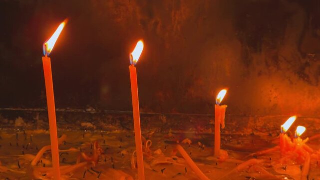 row of slim orange church candle burning with warm flame and dripping melted wax inside a dim sacred prayer space