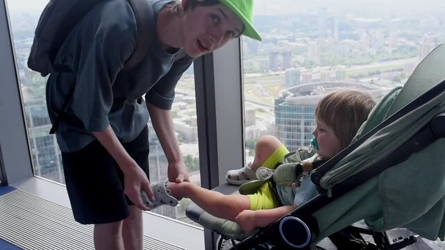 a  13-year-old teenage boy in a green cap putting on a sandal shoe on his 1-year-old baby brother's foot inside a stroller by a panoramic skyscraper window during a summer day