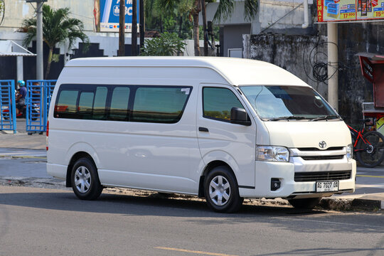 A white Toyota HiAce commercial commuter van parked on the side of a street with shop signs and buildings in the background.