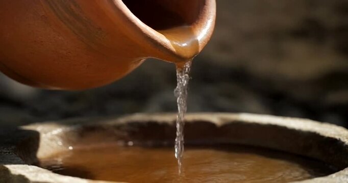 Close up of water pouring from ancient clay jug into stone basin outdoors.