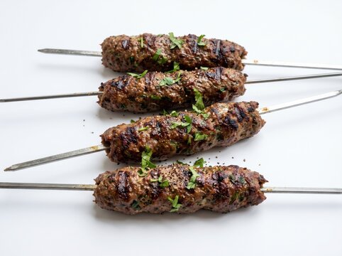 Four freshly grilled Turkish kofta kebabs on metal skewers garnished with green herbs, isolated on a bright white background, ideal for Middle Eastern restaurant menu design and food blogs.