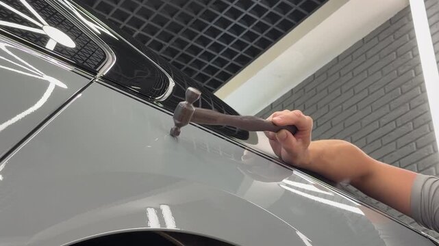 Mechanic hand fixing a car body damage using paintless dent repair PDR tools in a workshop. Close up of professional car detailing technician removing a dent with a special hammer compound. 