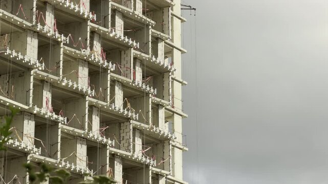 A concrete residential building stands mid-construction under a heavy, overcast sky. The empty window frames and raw materials create an overwhelming, bleak, and somber mood.