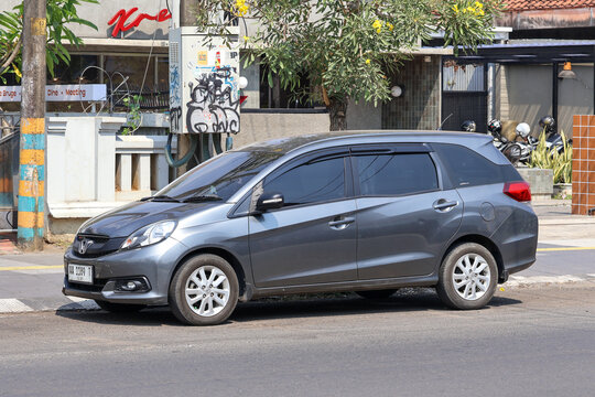 Side profile of a dark gray Honda Mobilio MPV parked on a street in Indonesia. The multi-purpose vehicle features window visors, dark tinted windows, and original alloy wheels on asphalt road.