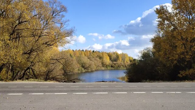 The river flows under an asphalt road with markings. Shrubs, birches and willows with yellow foliage grow on the banks. There are ripples on the water. Sunny autumn weather and blue sky with clouds