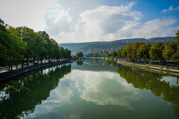 Kura River and iconic Bridge of Peace reflecting in Tbilisi, Georgia, with vibrant green trees. © Greggory Zzyzx