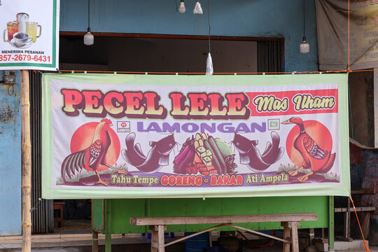 A traditional hand-painted fabric banner of a local indonesian street food stall, known as 'warung lamongan' or 'pecel lele', hanging in front of a modest eatery. the colorful banner.