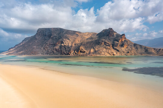 Aerial view of Detwah Lagoon with its turquoise waters, sandy beach, and rugged mountains under a cloudy sky in Qalansiyah wa Abd Al Kuri, Socotra, Yemen.