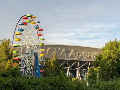View of the SKA Arena stadium building and a colorful classic Ferris wheel in a city amusement park surrounded by green summer trees under a blue sky.