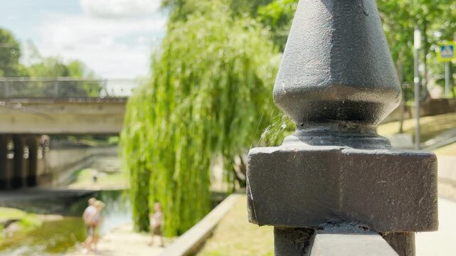 A black iron post stands by a sunny walkway. In the blurred background, carefree children play near a serene river. This warm summer day evokes a peaceful and nostalgic feeling.