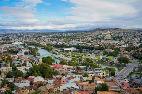 Panoramic view of Tbilisi, Georgia, with Mtkvari River, Bridge of Peace, and Sameba Cathedral under cloudy sky.