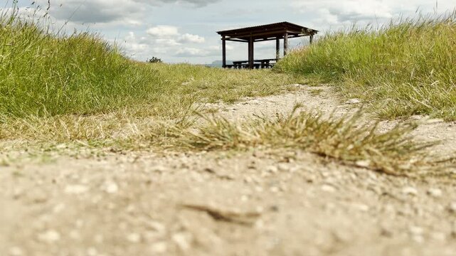 A lonely wooden gazebo stands quietly on top of a grassy hill. A dusty dirt path leads up to this peaceful shelter in the scenic mountains. The calm view brings a sense of pure relaxation.