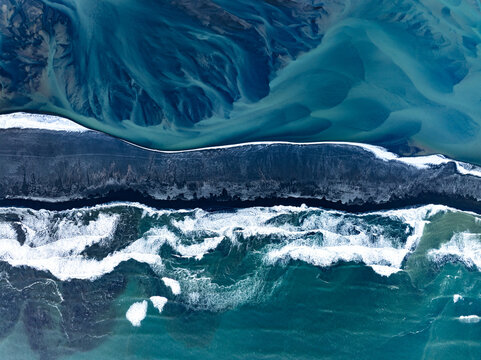 Aerial view of the dramatic Icelandic coastline where glacial river patterns meet the dark volcanic sand beach and the crashing waves of the Atlantic Ocean in Iceland.